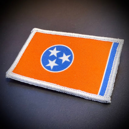 An embroidered rectangular patch of the Tennessee state flag, featuring an orange field, a blue circle with three white stars, and a blue vertical stripe, finished with a white stitched border, shown on a dark textured background.