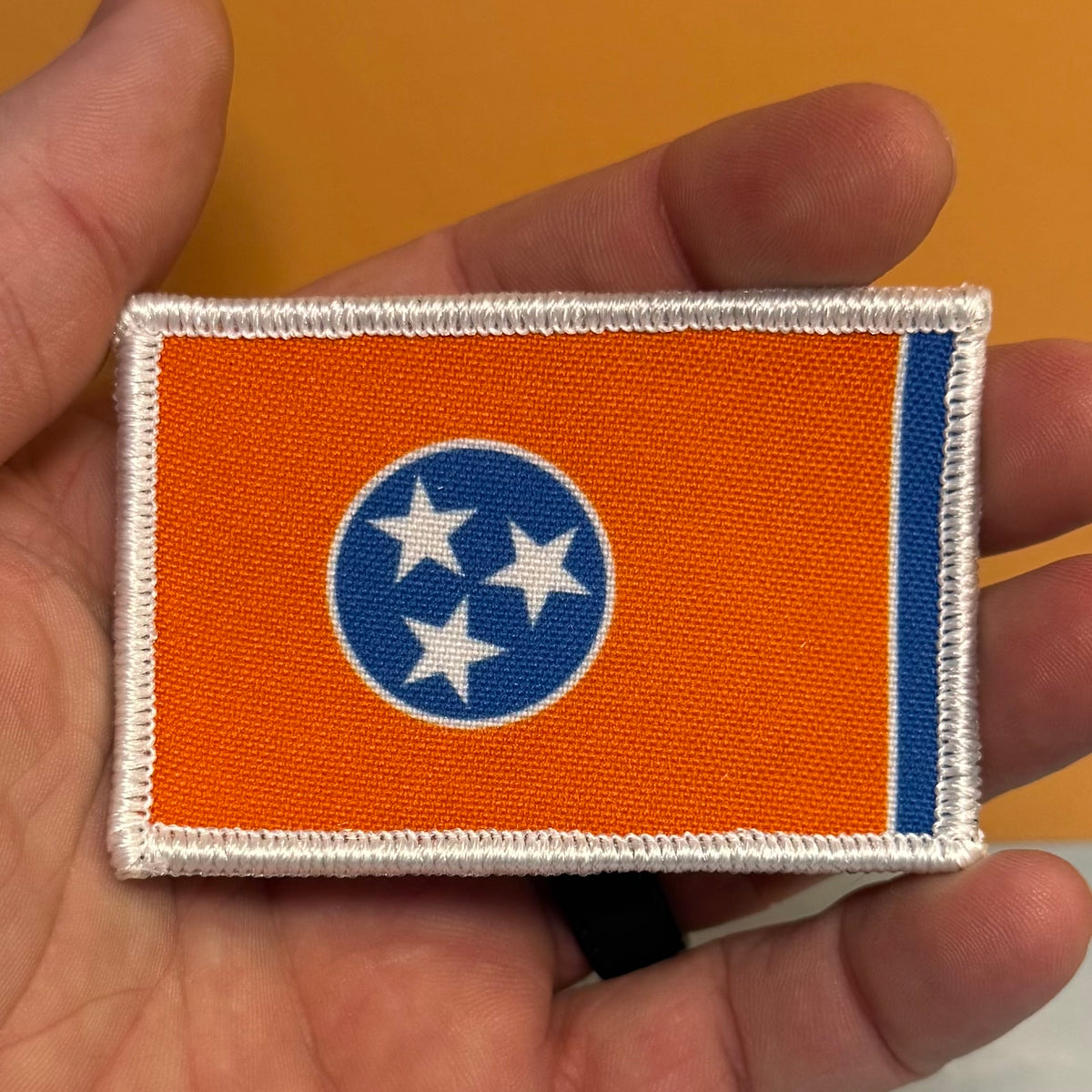 Tennessee Orange and Summitt Blue Flag printed canvas morale patch, of the Tennessee state flag held in a hand. The patch has an orange field, a blue circle with three white stars, and a blue vertical stripe on the fly end.
