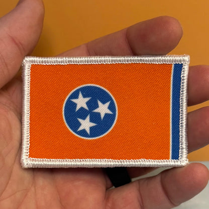 Tennessee Orange and Summitt Blue Flag printed canvas morale patch, of the Tennessee state flag held in a hand. The patch has an orange field, a blue circle with three white stars, and a blue vertical stripe on the fly end.