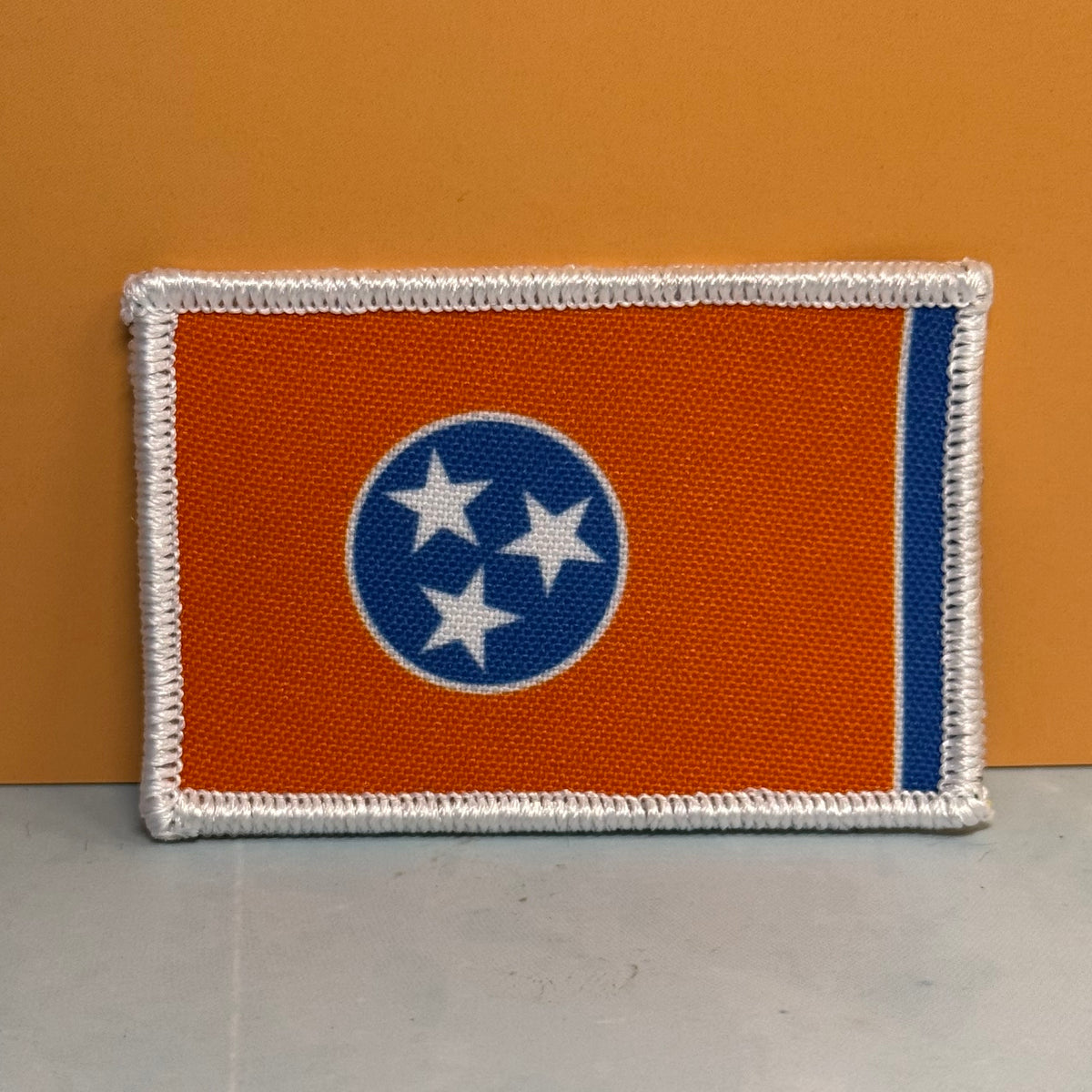 Tennessee Orange and Summitt Blue Flag printed canvas morale patch, of the Tennessee state flag, featuring an orange field, a blue circle with three white stars, and a vertical blue stripe, finished with a white merrowed.