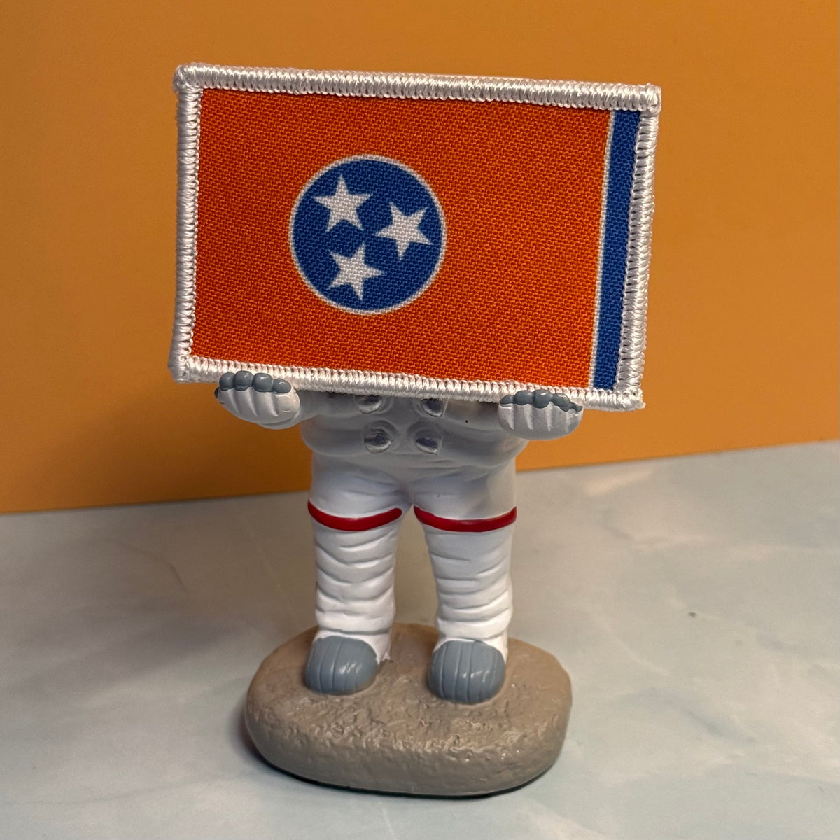 Tennessee Orange and Summitt Blue Flag printed canvas morale patch, a small astronaut figurine standing on a round gray base, holding a rectangular orange flag with a blue circle and three white stars. The figurine is dressed.