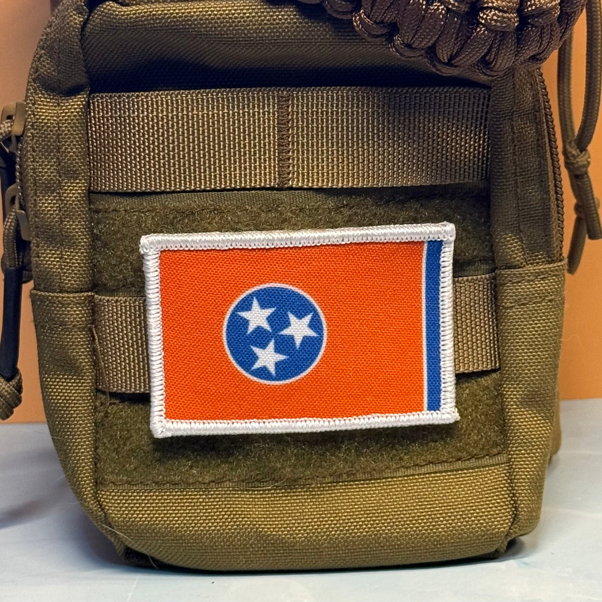 Tennessee Orange and Summitt Blue Flag printed canvas morale patch, an printed Tennessee state flag patch with a white border, orange field, blue circle, and three white stars, attached to the front of an olive drab tactical.