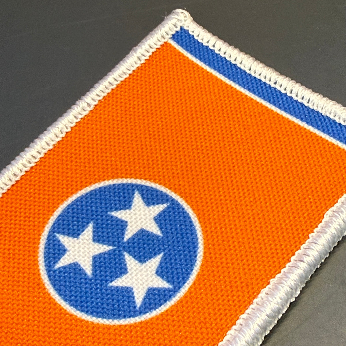 Tennessee Orange and Summitt Blue Flag printed canvas morale patch, of the Tennessee state flag, featuring an orange field, a blue circle with three white stars, and a white border with blue trim, shown on a dark gray background.