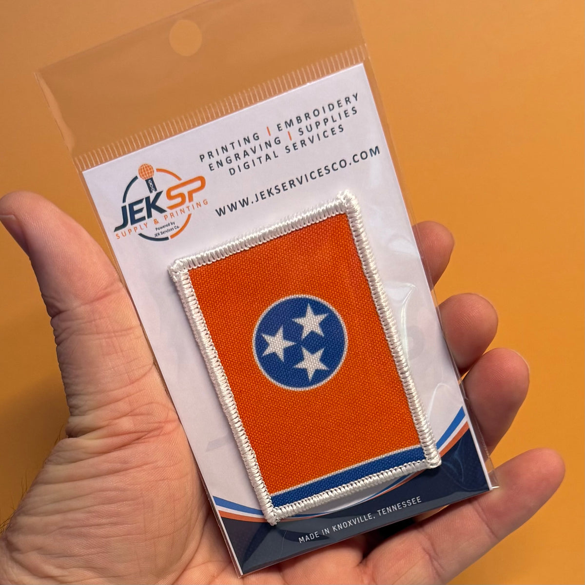 Tennessee Orange and Summitt Blue Flag printed canvas morale patch, a hand holding a small printed Tennessee state flag patch on a branded backing card. The patch is orange with a blue circle and white stars, bordered in.