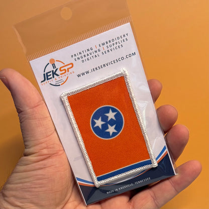 Tennessee Orange and Summitt Blue Flag printed canvas morale patch, a hand holding a small printed Tennessee state flag patch on a branded backing card. The patch is orange with a blue circle and white stars, bordered in.