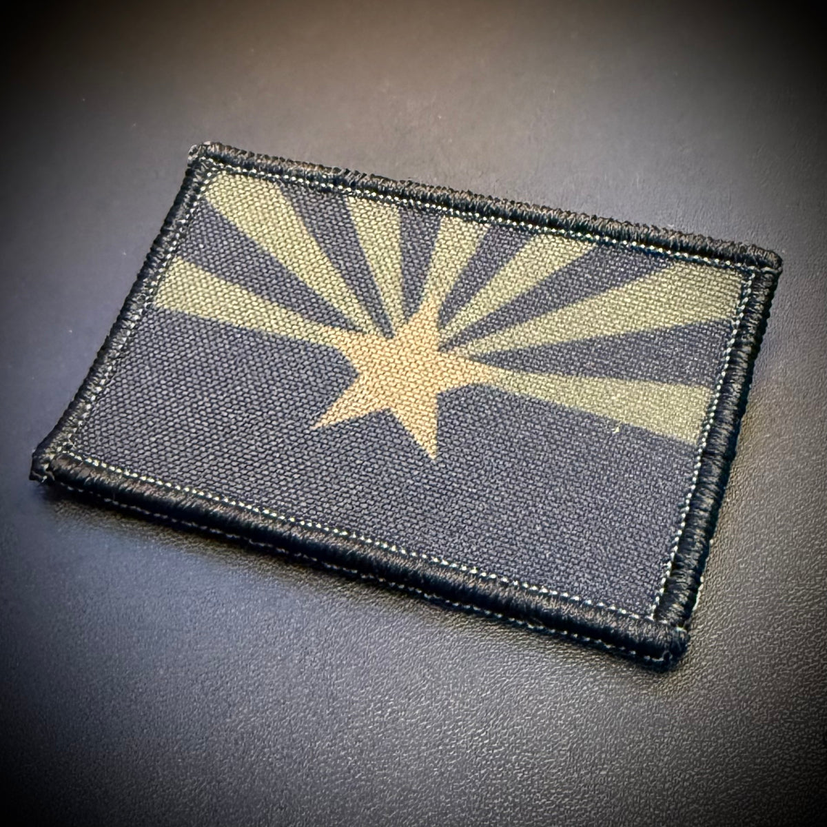 A rectangular tactical patch featuring the Arizona state flag design with a tan star and rising sun rays on a dark charcoal background, finished with a black stitched border, shown on a dark textured surface.