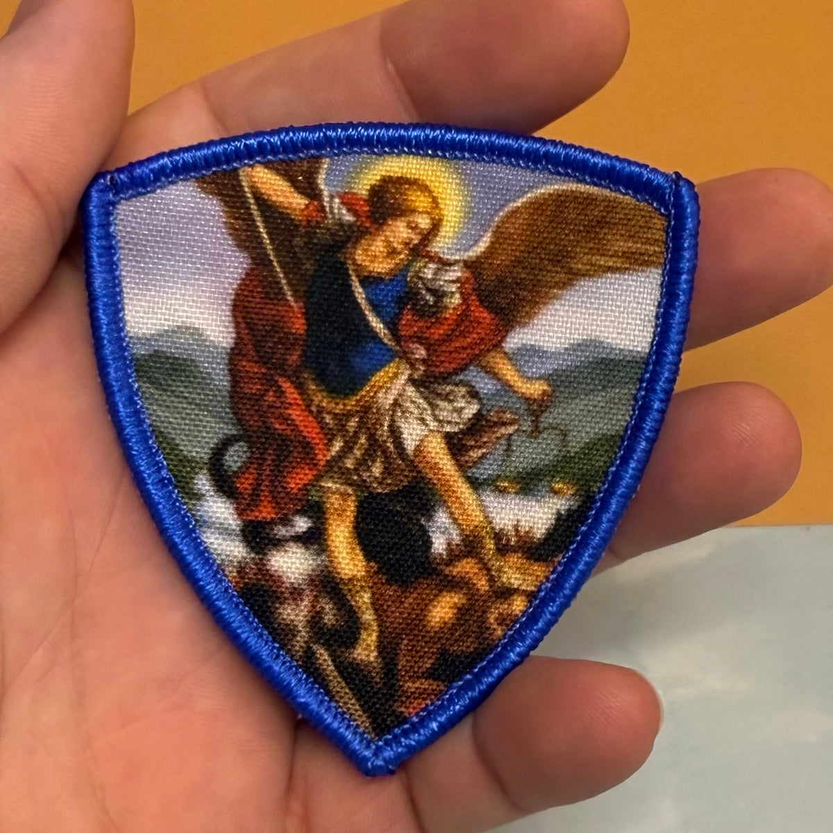 Saint Michael Shield printed canvas morale patch, a shield-shaped printed patch featuring a religious scene of an angel with wings and a sword, held in a person's hand against a yellow and white background.