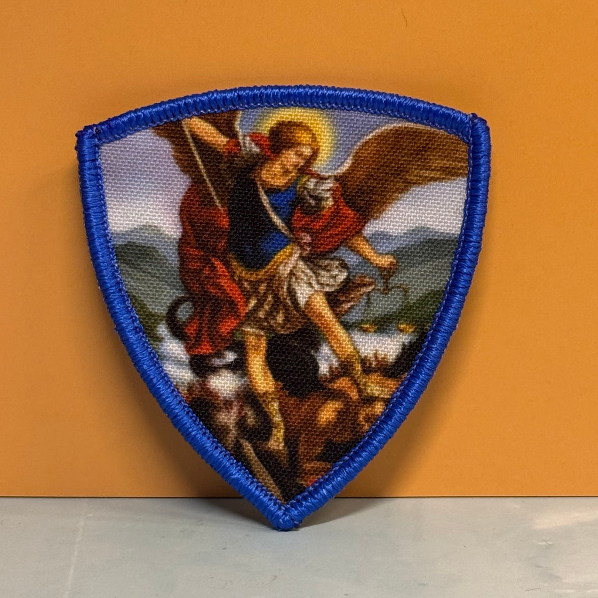 Saint Michael Shield printed canvas morale patch, a shield-shaped printed patch featuring a religious scene of an angel with wings and a staff, set against a blue stitched border, displayed against a solid orange background.