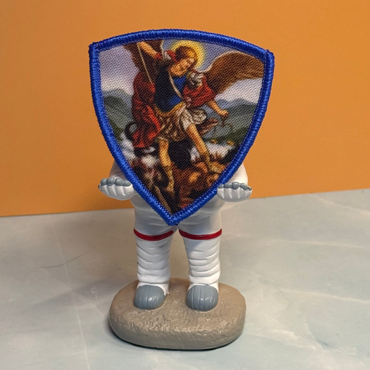 Saint Michael Shield printed canvas morale patch, a small figurine or doll with a shield-shaped printed patch on its head, standing on a round base against an orange and gray background.