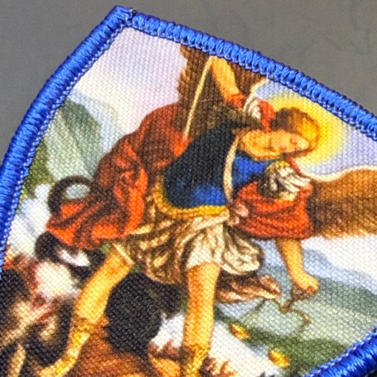 Saint Michael Shield printed canvas morale patch, a close-up of a fabric patch featuring a classical religious illustration of an angel with wings and a halo, wearing a red robe and blue tunic, bordered by a blue stitched edge.