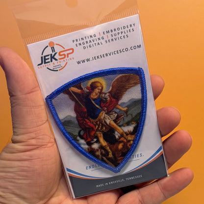 Saint Michael Shield printed canvas morale patch, a hand holds a packaged printed patch featuring a blue-bordered shield with a religious scene. The backing card shows the Jekspaint logo and text for printing, embroidery.