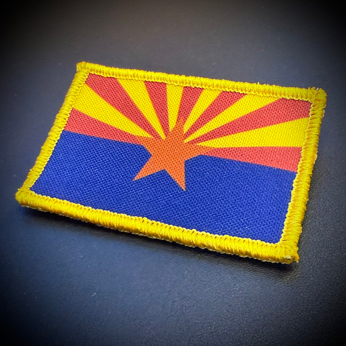 An embroidered patch of the Arizona state flag with a yellow border, featuring red and yellow rays, a central orange star, and a blue lower section, displayed on a dark textured background.