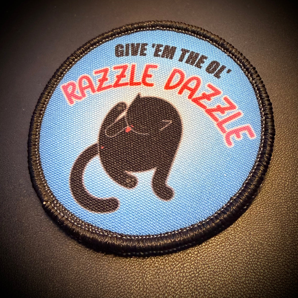 A circular embroidered patch featuring a black cat silhouette on a light blue background, with the text 'GIVE 'EM THE OL" RAZZLE DAZZLE' in black and red lettering, shown on a dark textured surface.