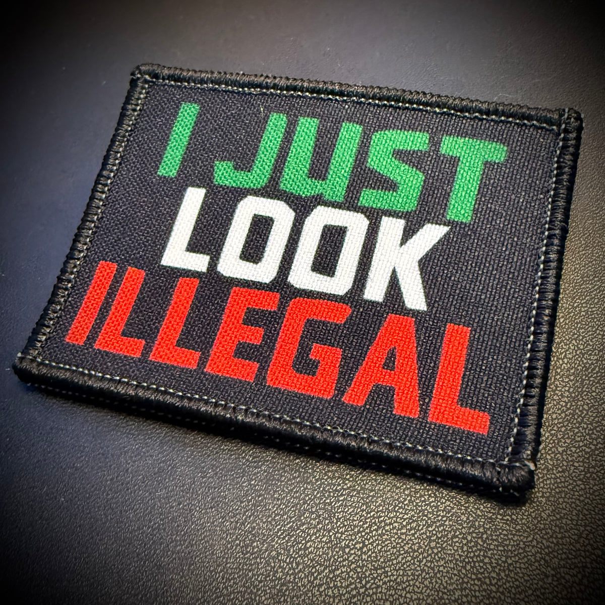 A rectangular embroidered patch with the text 'I JUST LOOK ILLEGAL' in green, white, and red lettering on a black background, placed on a dark textured surface.