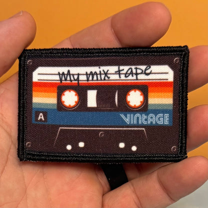 A rectangular embroidered patch shaped like a cassette tape held in a person's hand. The patch reads 'My mix tape' and 'VINTAGE' with retro orange, yellow, cream, and blue stripes on a dark background.