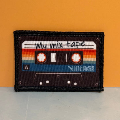 A rectangular embroidered patch shaped like a vintage cassette tape, with the words 'my mix tape' and 'VINTAGE' on a retro striped background, shown against an orange wall and light gray surface.