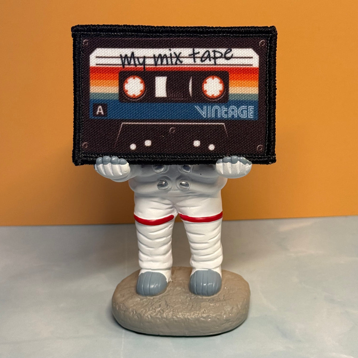 A small astronaut figurine standing on a round base, holding a large retro cassette tape patch over its head. The patch reads 'My mix tape' and 'Vintage' with rainbow stripes.