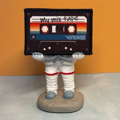A small astronaut figurine standing on a round base, holding a large retro cassette tape patch over its head. The patch reads 'My mix tape' and 'Vintage' with rainbow stripes.