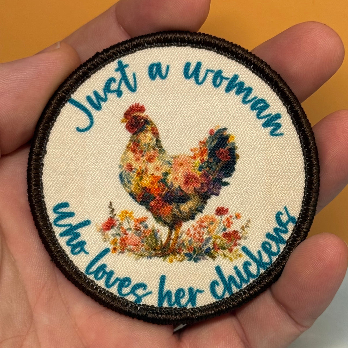 A round embroidered patch held in a hand, featuring a colorful floral rooster and the text 'Just a woman who loves her chickens' in blue script on a cream background with a dark brown border.