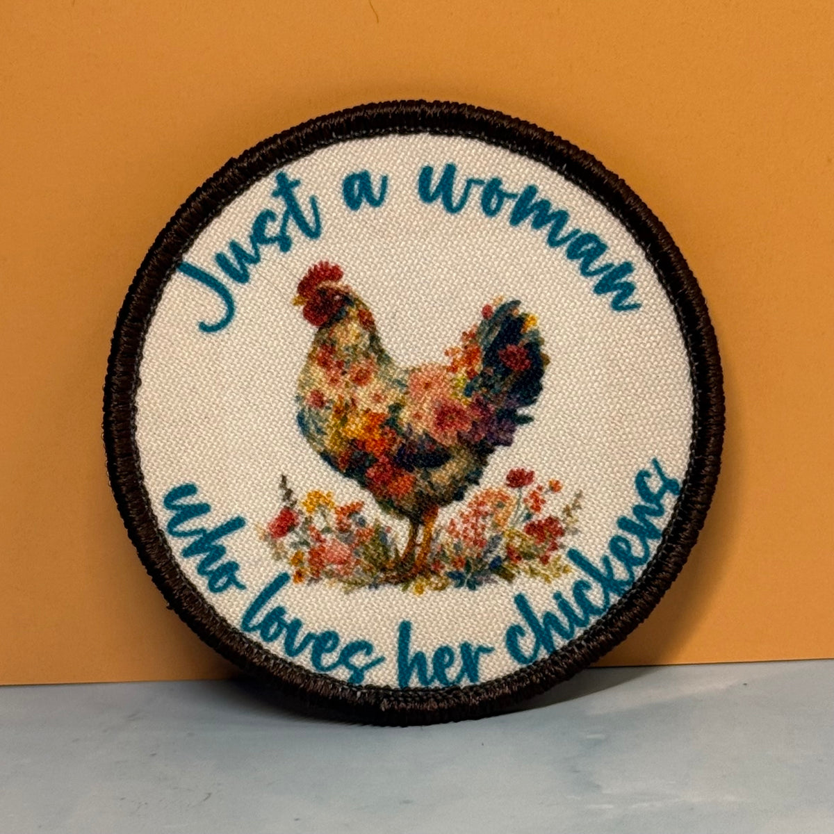 A round embroidered patch with a dark brown stitched border, featuring a colorful floral rooster in the center and blue embroidered text that reads 'Just a woman who loves her chickens' on a cream fabric background, set against a mustard yellow wall.
