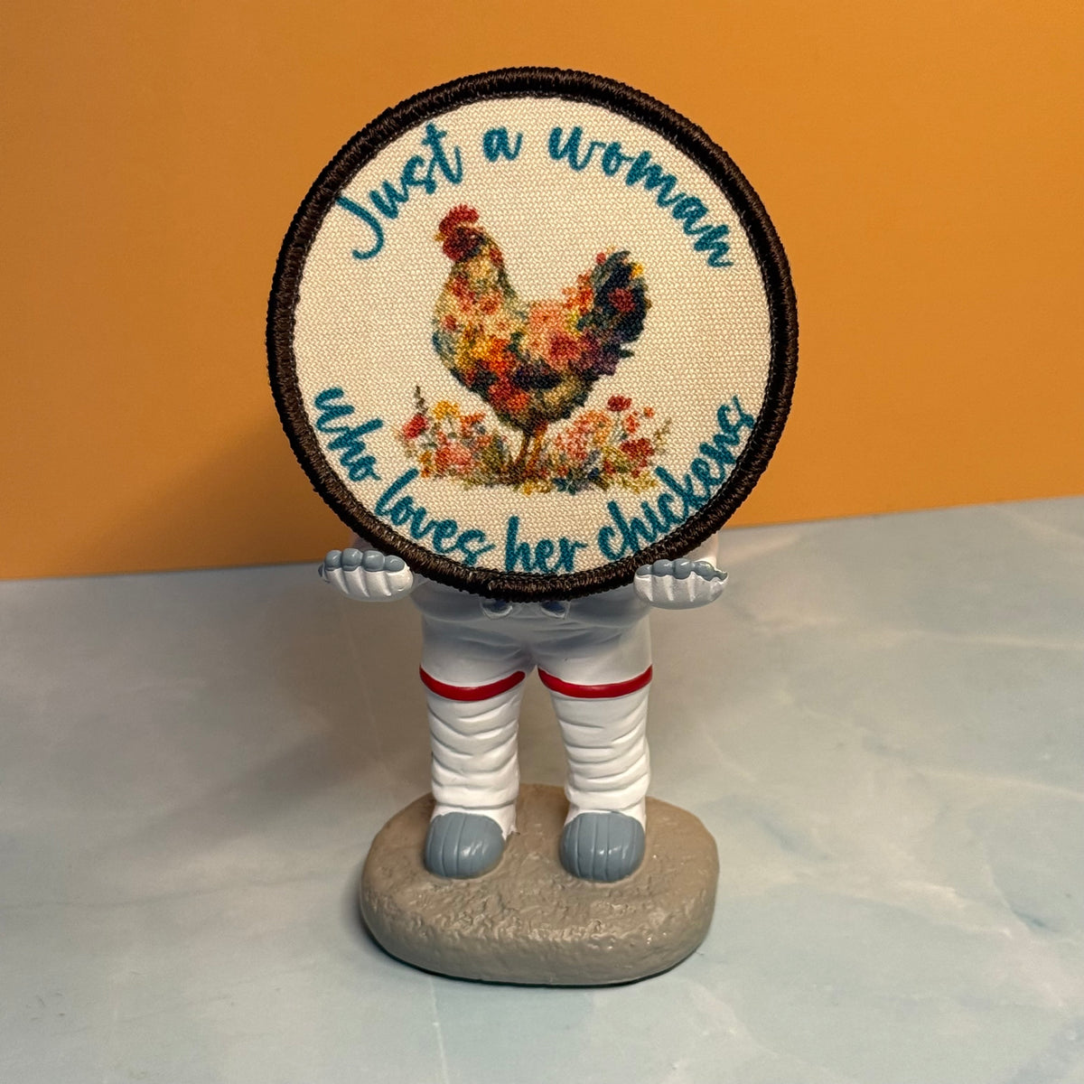 A round embroidered patch featuring a colorful floral rooster and the text 'Just a woman who loves her chickens' on a beige fabric background with a black stitched border. The patch is held by a small figurine with white pants, gray feet, and red accents, set against an orange and gray background.