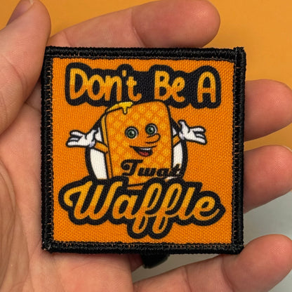 A square embroidered patch held in a hand, featuring an orange background with the text 'Don't Be A Twat Waffle' in yellow and black, and a cartoon waffle character in the center.