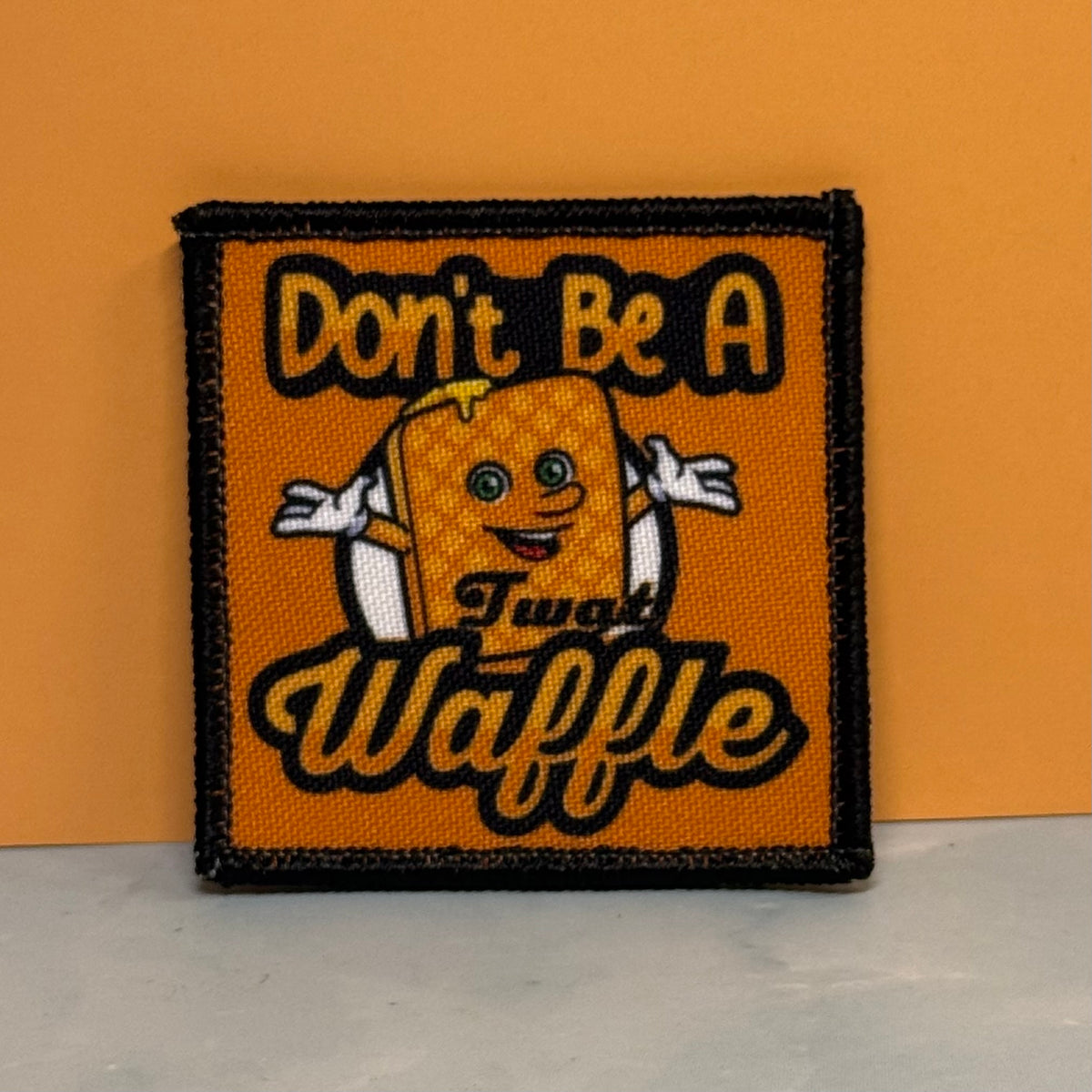 An embroidered square patch featuring a cartoon waffle character with the text 'Don't Be A Twat Waffle' on an orange background with a black stitched border, set against an orange wall and light gray floor.
