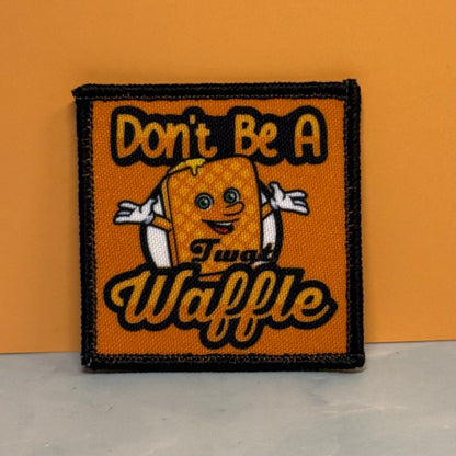 An embroidered square patch featuring a cartoon waffle character with the text 'Don't Be A Twat Waffle' on an orange background with a black stitched border, set against an orange wall and light gray floor.
