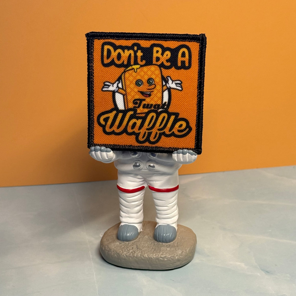 A small astronaut figurine standing on a gray base, holding a square orange patch that reads “Don’t Be A Tug Waffle” with a cartoon waffle character. The figurine is dressed in a white spacesuit with red bands and gray boots, set against an orange wall and light gray surface.