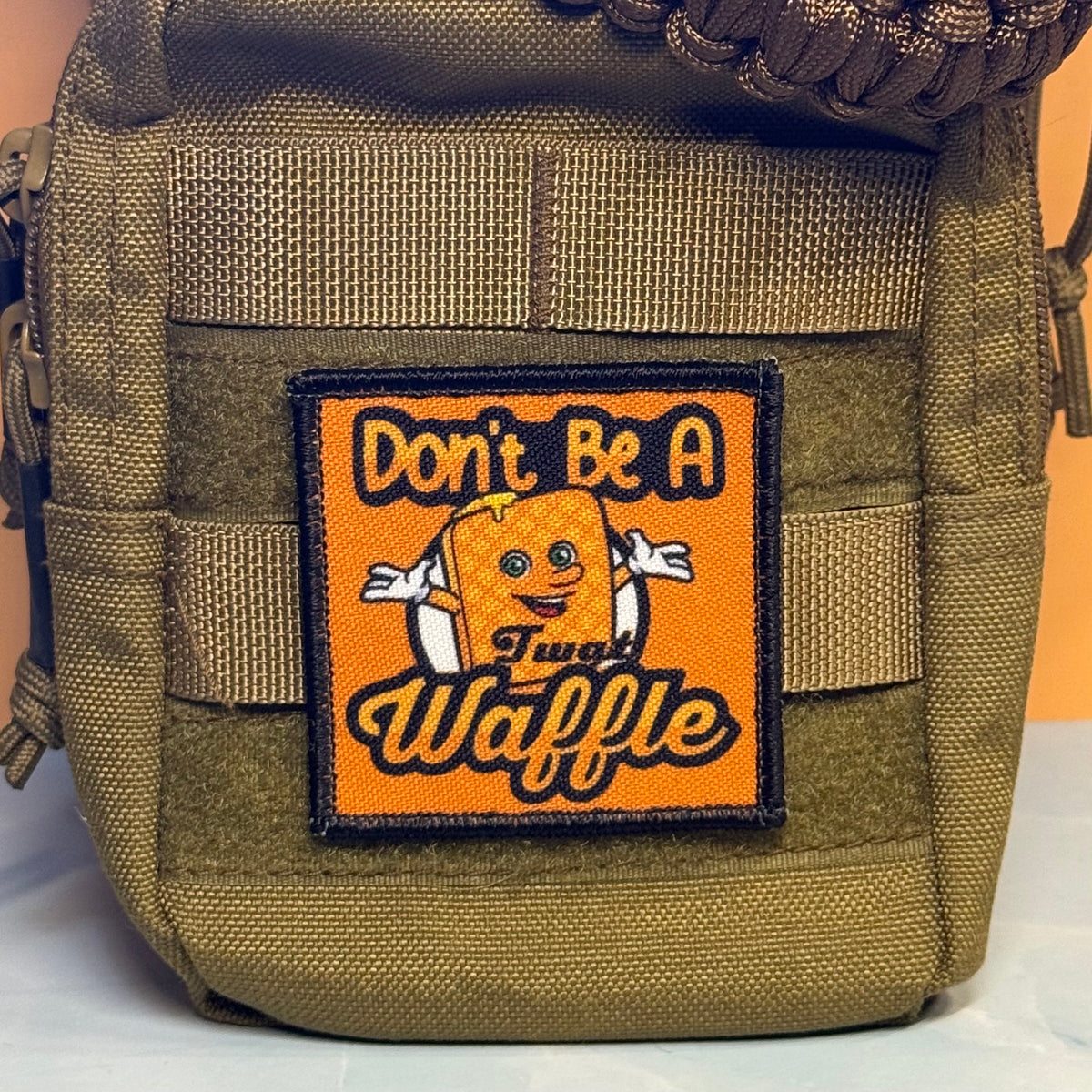 An olive green tactical pouch featuring a square orange patch with a cartoon waffle and the text 'Don't Be A Sweet Waffle'.