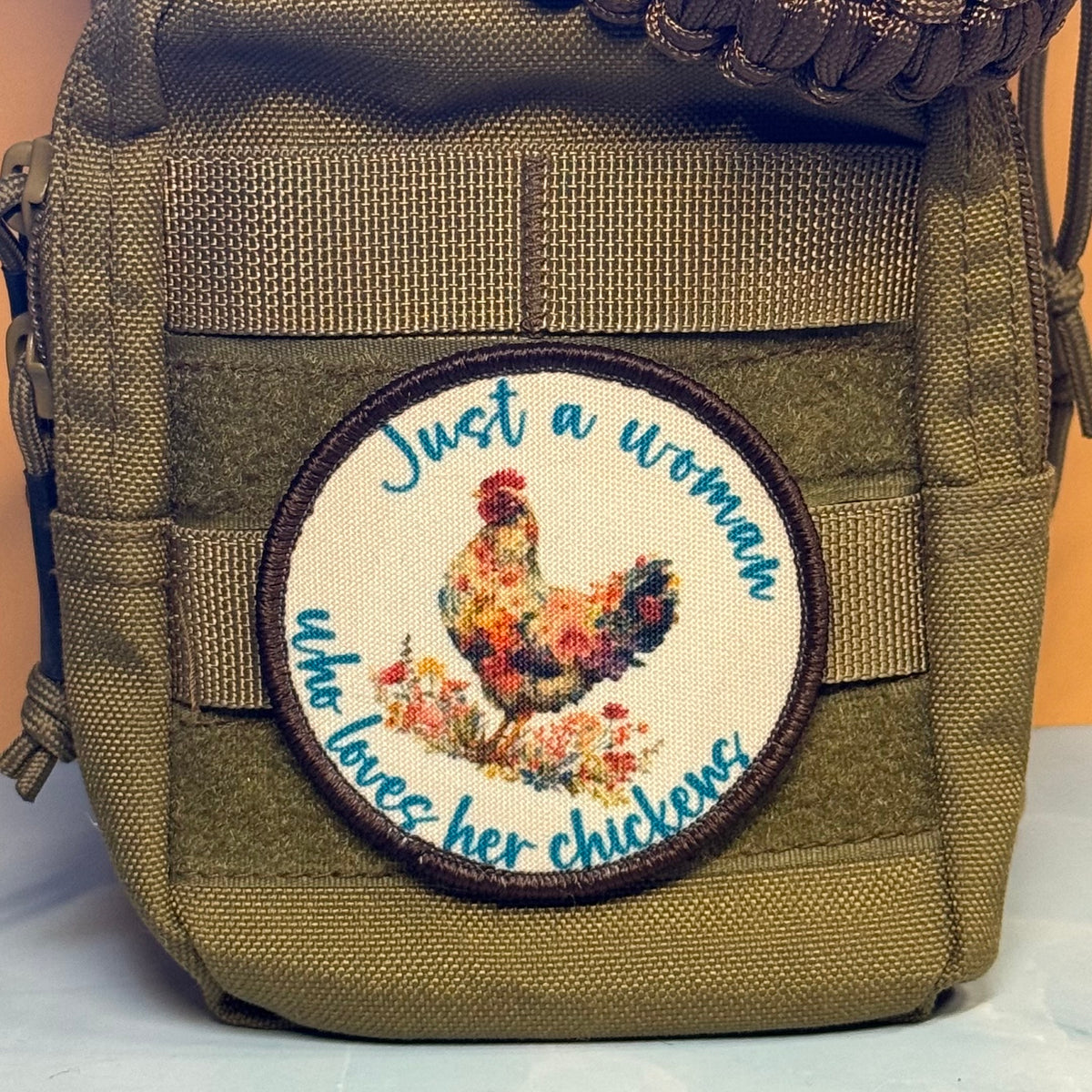 An olive green tactical pouch featuring a circular embroidered patch with a floral rooster and the text 'Just a woman who loves her chickens'.