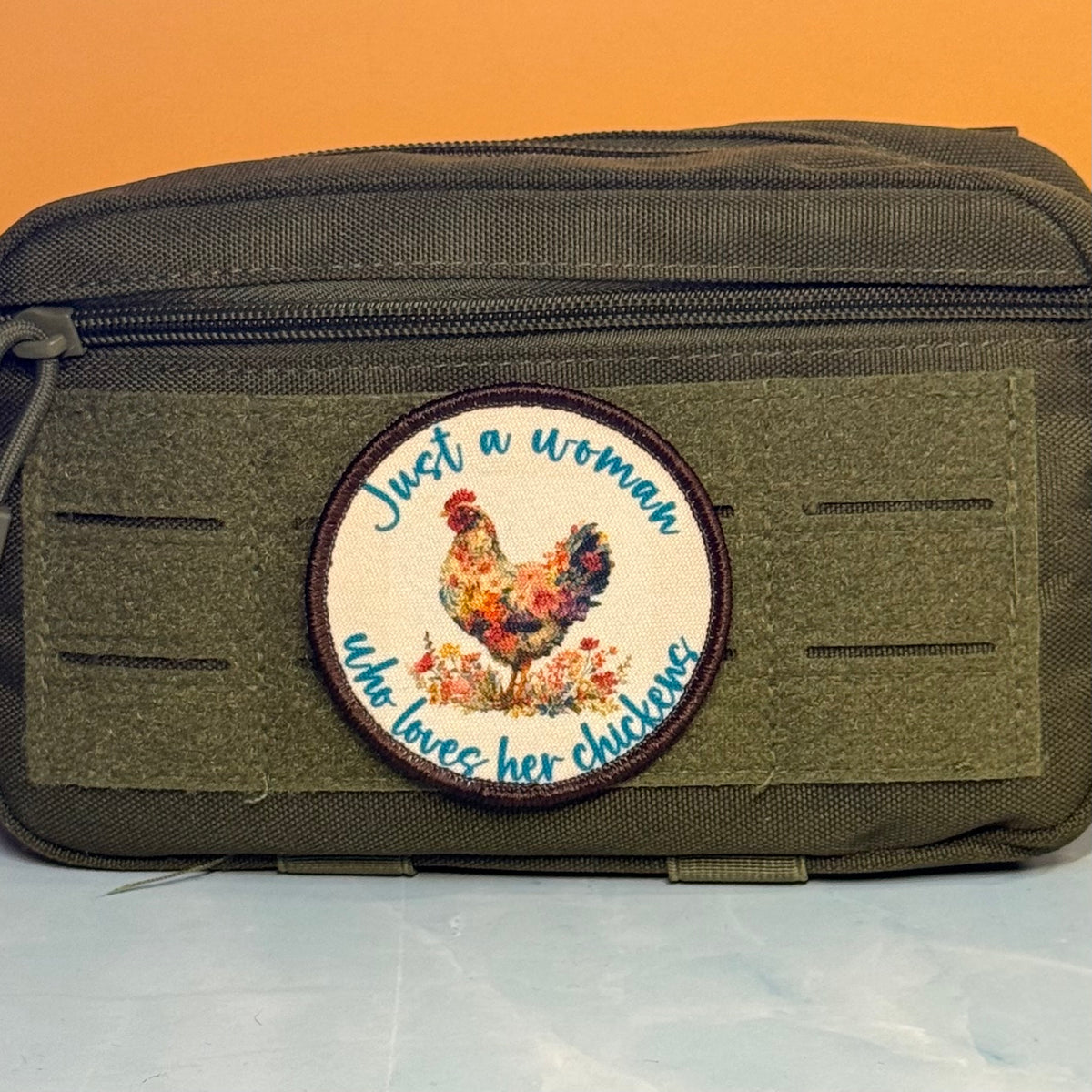 An olive green tactical pouch with a circular embroidered patch featuring a rooster and the text 'Just a woman who loves her chickens' on a white background.