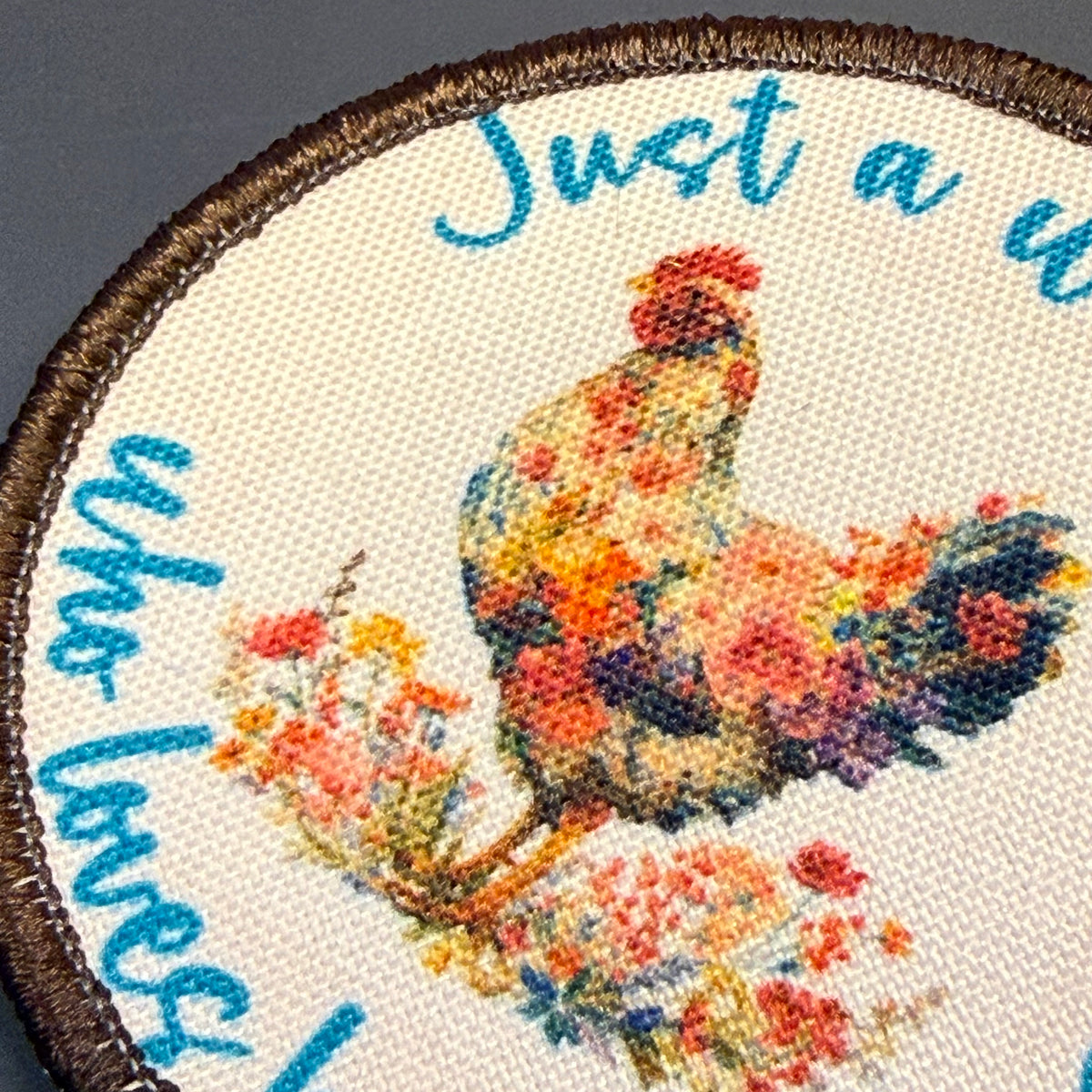 A close-up of a circular embroidered patch featuring a colorful floral rooster on an off-white fabric background, surrounded by blue cursive text and a brown stitched border.