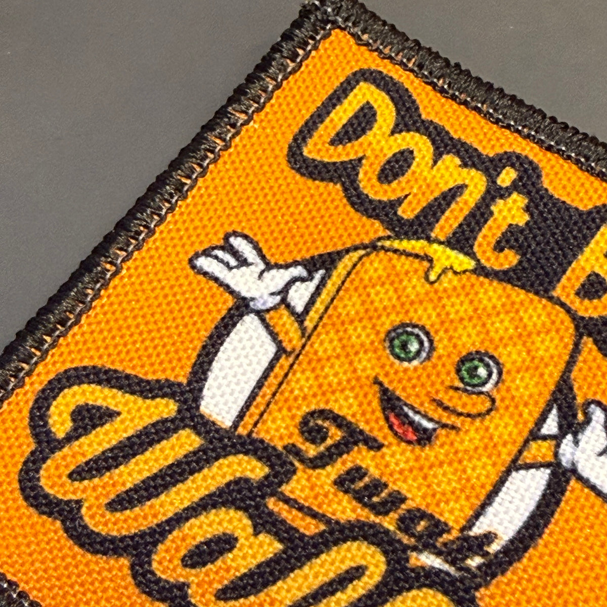 A close-up of a woven embroidered patch featuring an orange cartoon soda can character with the text 'Don't Be Twat Wan' in stylized lettering, on a dark gray background.