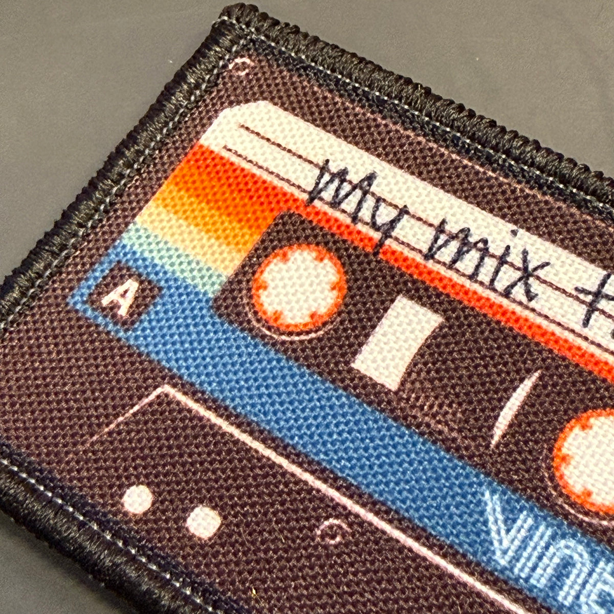 A close-up of a rectangular embroidered patch shaped like a cassette tape, featuring a brown body with blue, orange, and white graphic details and the handwritten text 'my mix +' across the top.