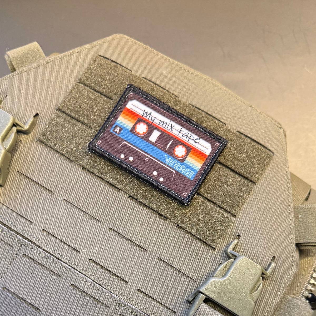 A rectangular tactical patch shaped like a cassette tape, featuring retro orange, red, blue, and white stripes with the text 'My Mix Tape' and 'Vintage', displayed on tan MOLLE webbing of a tactical vest.