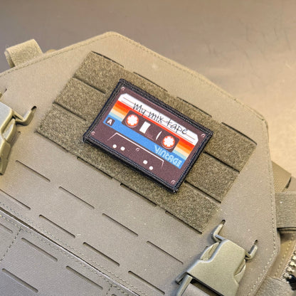 A rectangular tactical patch shaped like a cassette tape, featuring retro orange, red, blue, and white stripes with the text 'My Mix Tape' and 'Vintage', displayed on tan MOLLE webbing of a tactical vest.
