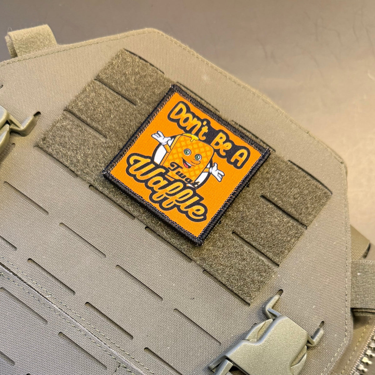 A rectangular embroidered patch with a yellow-orange background and a cartoon orange character, reading 'Don't Be A Wannabe,' attached to tan tactical webbing on a beige fabric pouch.