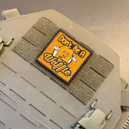 A rectangular embroidered patch with a yellow-orange background and a cartoon orange character, reading 'Don't Be A Wannabe,' attached to tan tactical webbing on a beige fabric pouch.