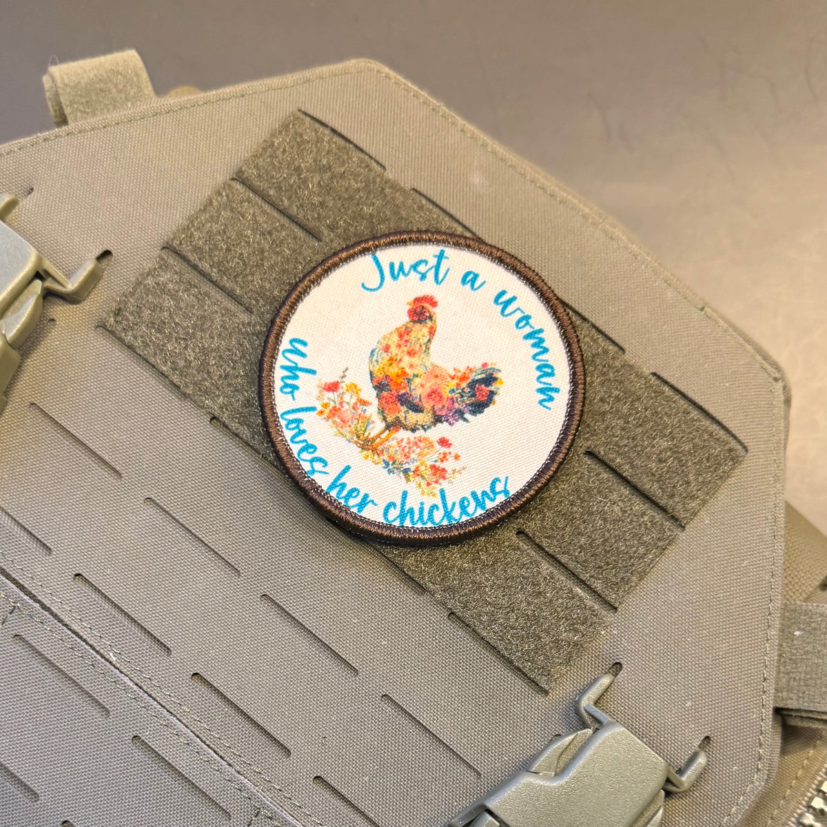 A circular embroidered patch featuring a colorful rooster and the text 'Just a woman who raises her chickens' in blue script, shown attached to a gray tactical vest with velcro and buckles.