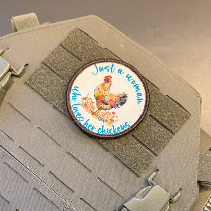A circular embroidered patch featuring a colorful rooster and the text 'Just a woman who raises her chickens' in blue script, shown attached to a gray tactical vest with velcro and buckles.