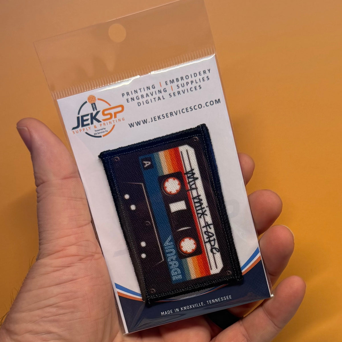 A hand holds a packaged embroidered patch shaped like a vintage cassette tape. The patch has a dark navy background with orange, yellow, red, and white striped details, black tape reels, and white text reading 'VINTAGE' and 'Misty Mayfield.