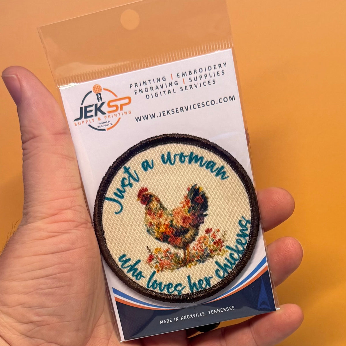 A hand holds a packaged round embroidered patch that reads 'Just a woman who loves her chickens' with a colorful chicken illustration. The patch is mounted on a white backing card with the JEKSP logo and text, against a solid orange background.