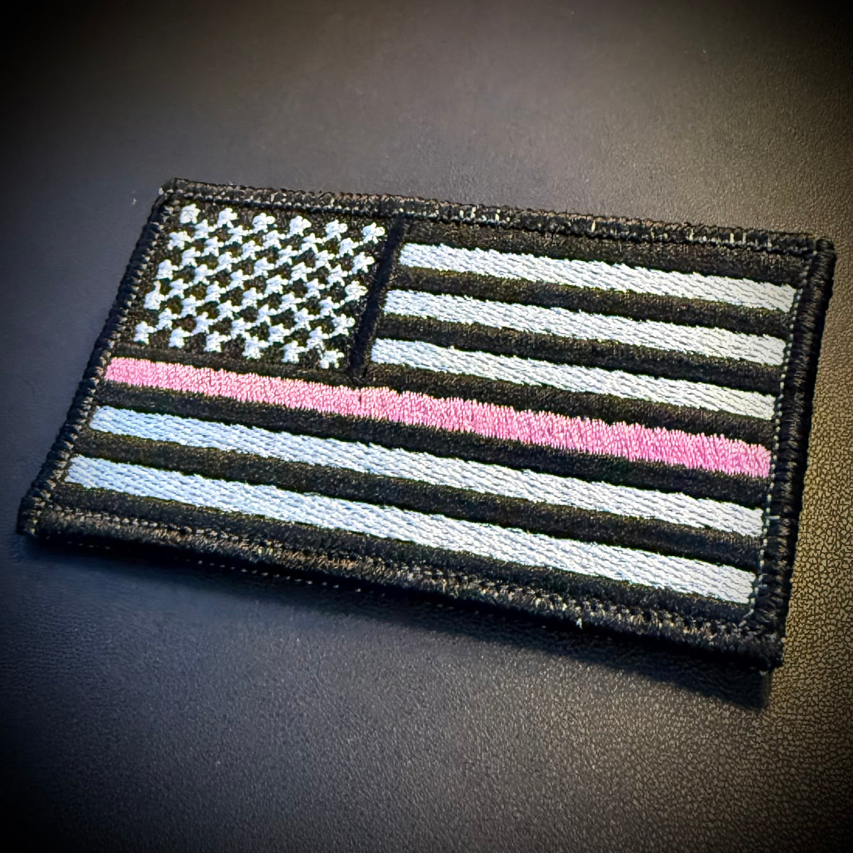 A rectangular embroidered American flag patch with a thin pink line, shown on a dark textured surface. The flag design uses black, white, and pink embroidery.