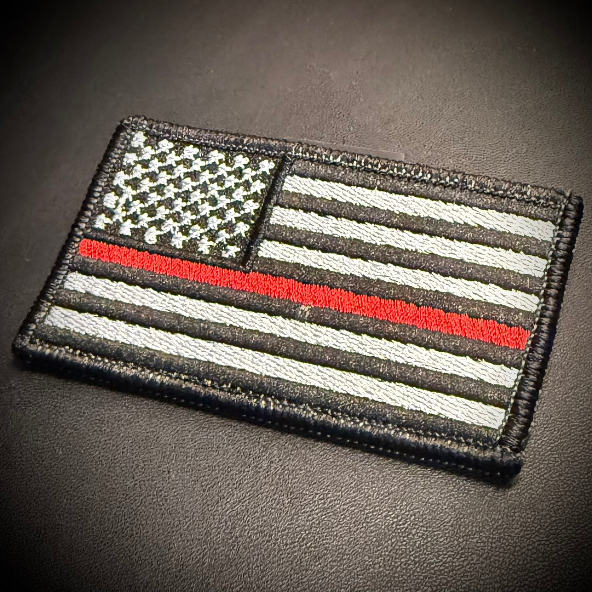 A rectangular embroidered patch featuring a black-and-white American flag with a thin red line, photographed on a dark textured surface.