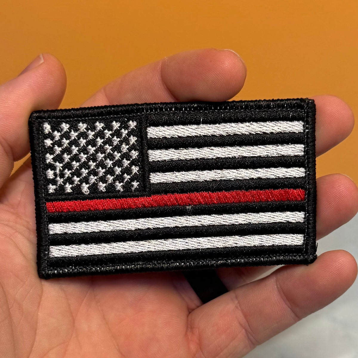 Thin Red Line American Flag Firefighter embroidered morale patch: hand holding a rectangular embroidered patch featuring a black-and-white American flag design with a single red stripe across the center, against a warm orange background.