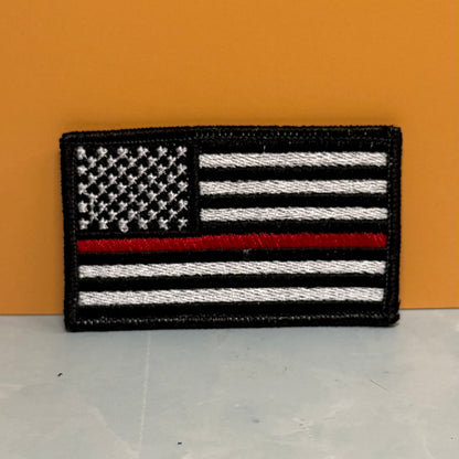 Thin Red Line American Flag Firefighter embroidered morale patch: featuring a black-and-white American flag with a single red horizontal stripe, photographed against an orange background and light gray surface.