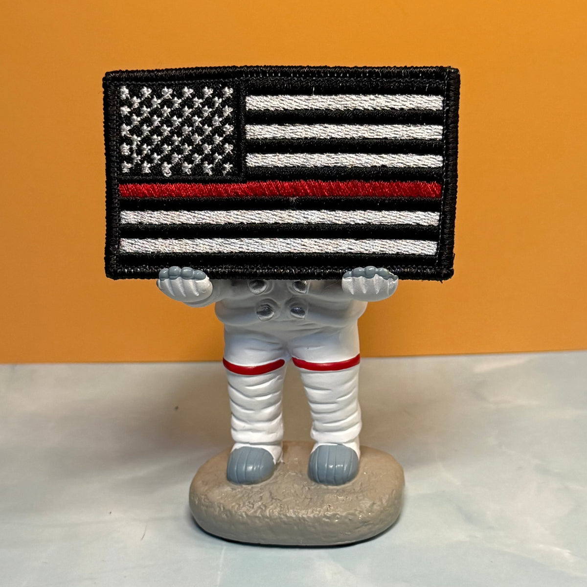 Thin Red Line American Flag Firefighter embroidered morale patch: astronaut figurine holding a black, white, and red embroidered American flag patch over its head, set against a solid orange background.