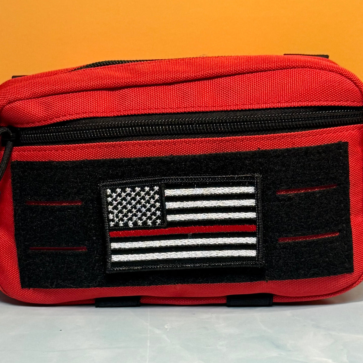 Thin Red Line American Flag Firefighter embroidered morale patch: red canvas utility pouch with a black zipper and a black front patch featuring a black-and-white American flag with a single red stripe, set against a solid orange background.