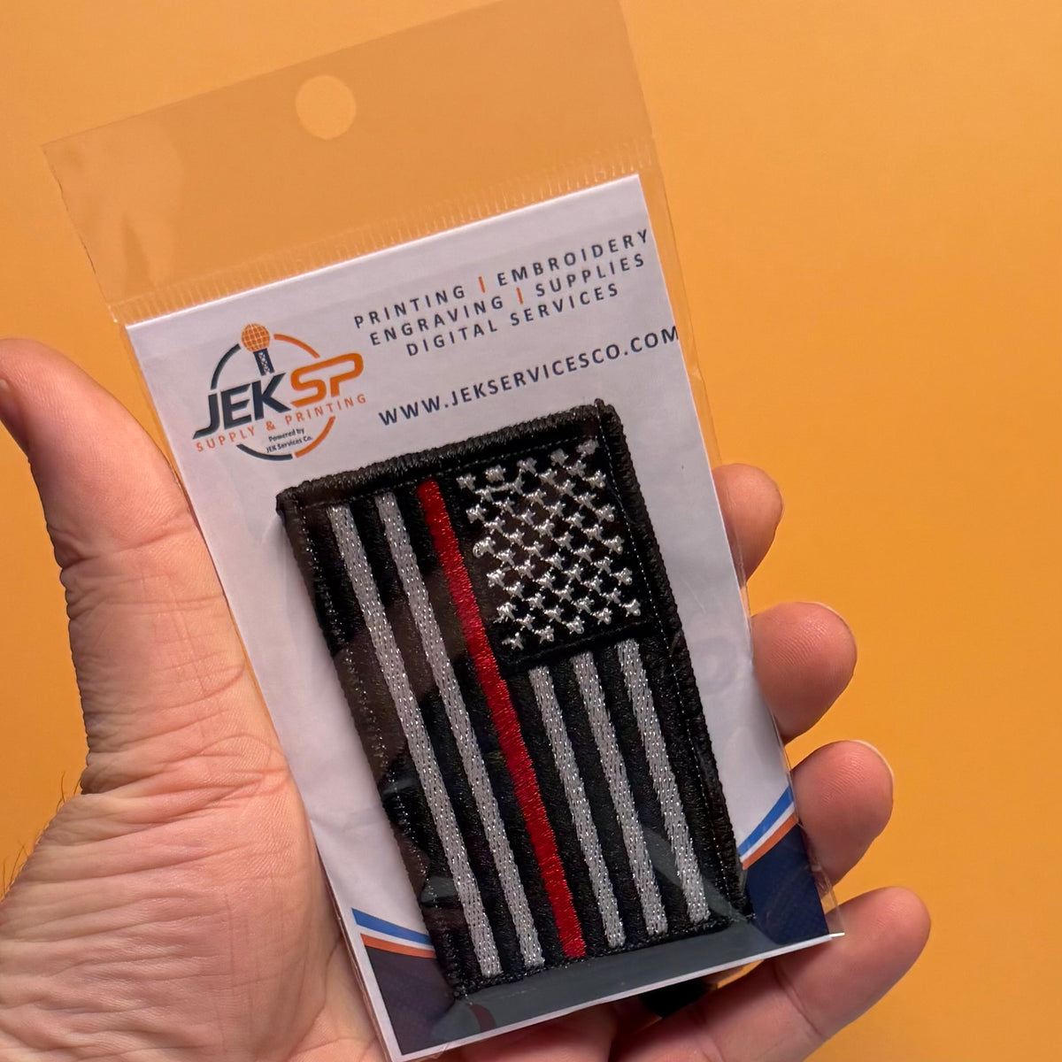 Thin Red Line American Flag Firefighter embroidered morale patch: hand holding a packaged embroidered patch featuring a black-and-white American flag design with a single red stripe, on a white backing card against an orange background.